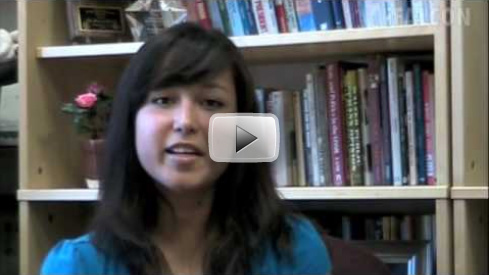 A video still of a female student sitting in front of a bookcase.