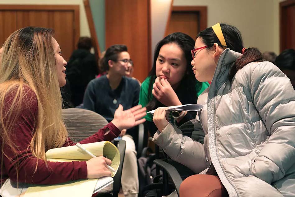 A group of young Asian American students sitting in a circle and engaging in an active discussion during an indoor meeting or workshop.