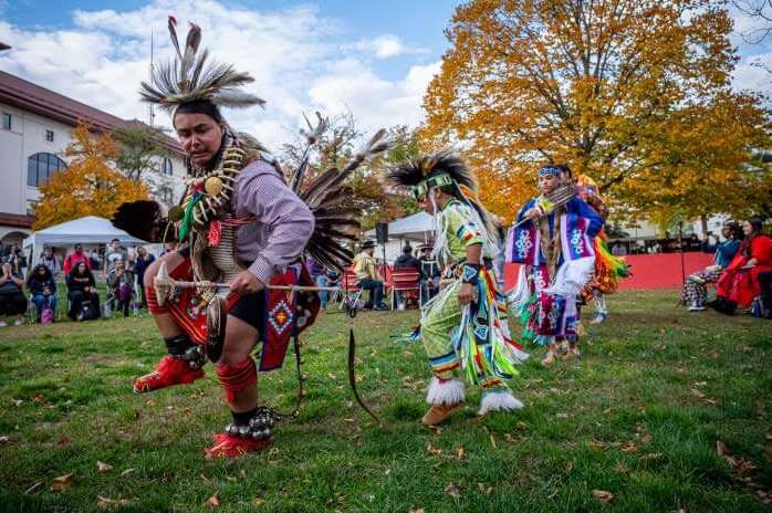 Native American dancers in traditional regalia performing outdoors on a grassy field during a community event or celebration.