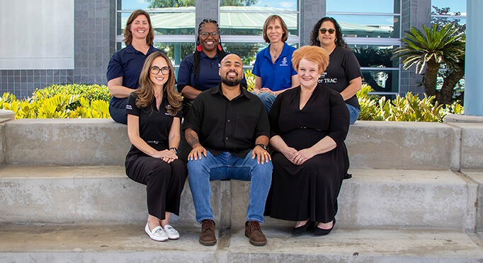 A group portrait of seven people smiling while posing outdoors on concrete steps in front of a building with large glass windows.