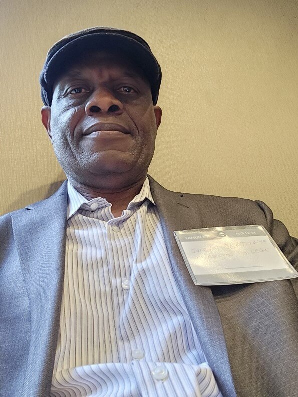 A man (Sunday Obazuaye) faces the camera, wearin gray tweed coat and hat.