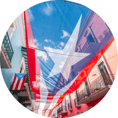 Circular cropped low-angle view of a translucent Puerto Rican flag canopy stretched across a narrow street framed by colorful historic buildings in Old San Juan.