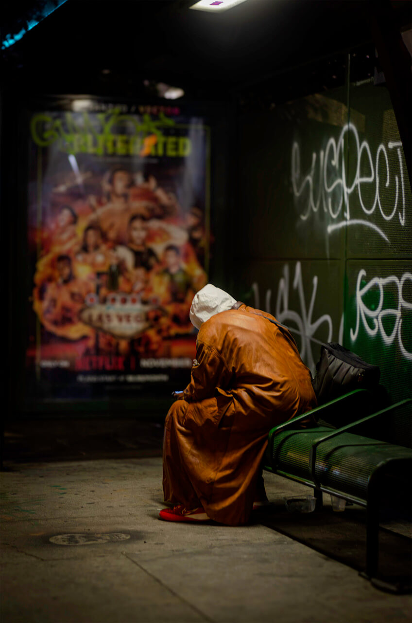 "Back of figure with orange-brown coat and white hood at green bus stop.
