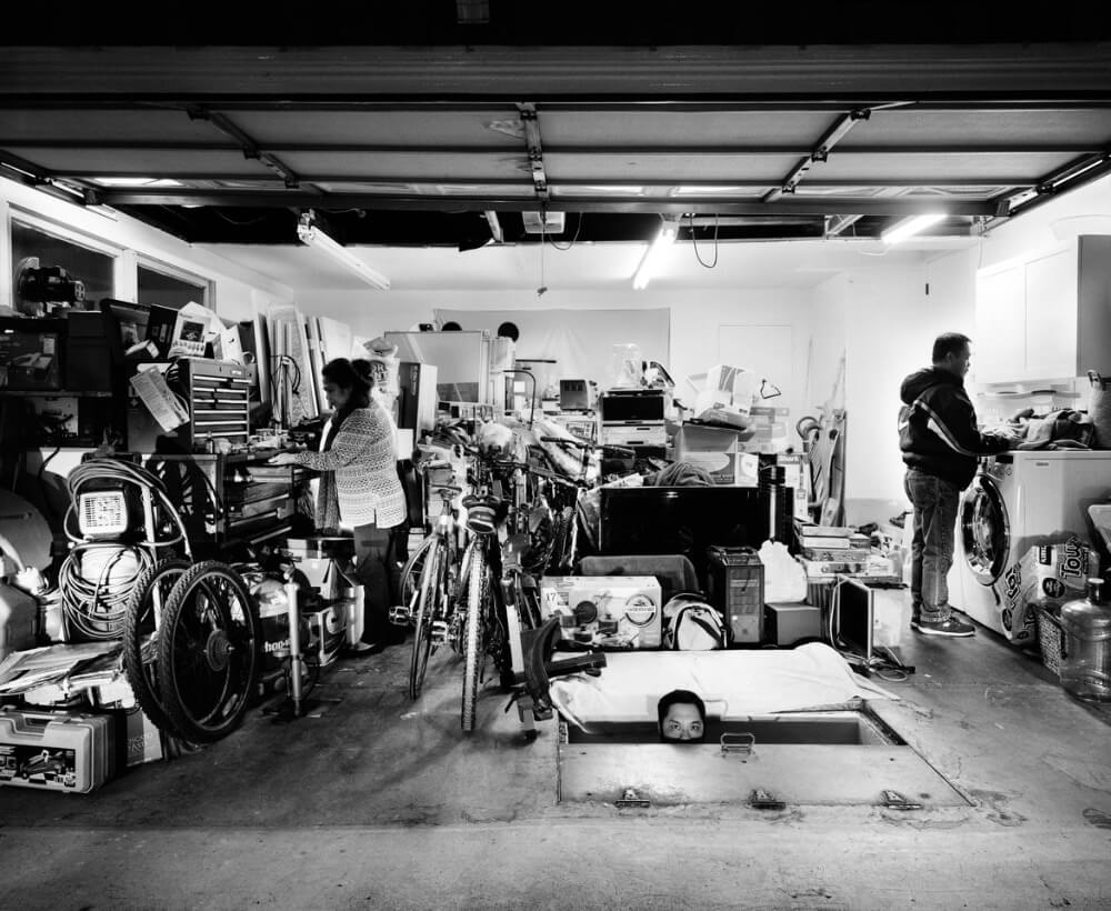 Man and woman inside cluttered garage and male head peeking from door on floor.