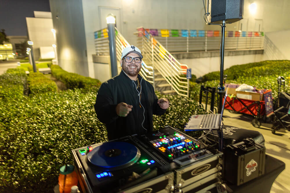 Smiling DJ in white cap and glasses at turntables and mixer outdoors at night, with papel picado on stairs behind.