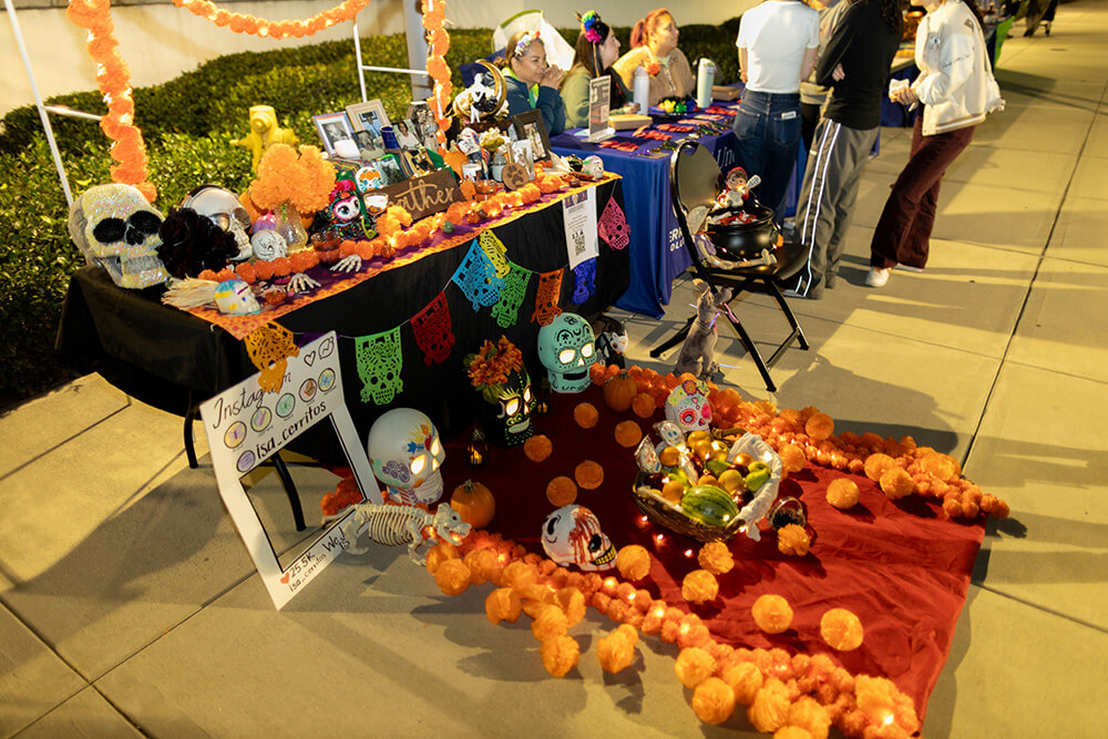 Día de los Muertos altar with sugar skulls, marigolds, photos, papel picado, and fruit basket on red cloth at outdoor event.