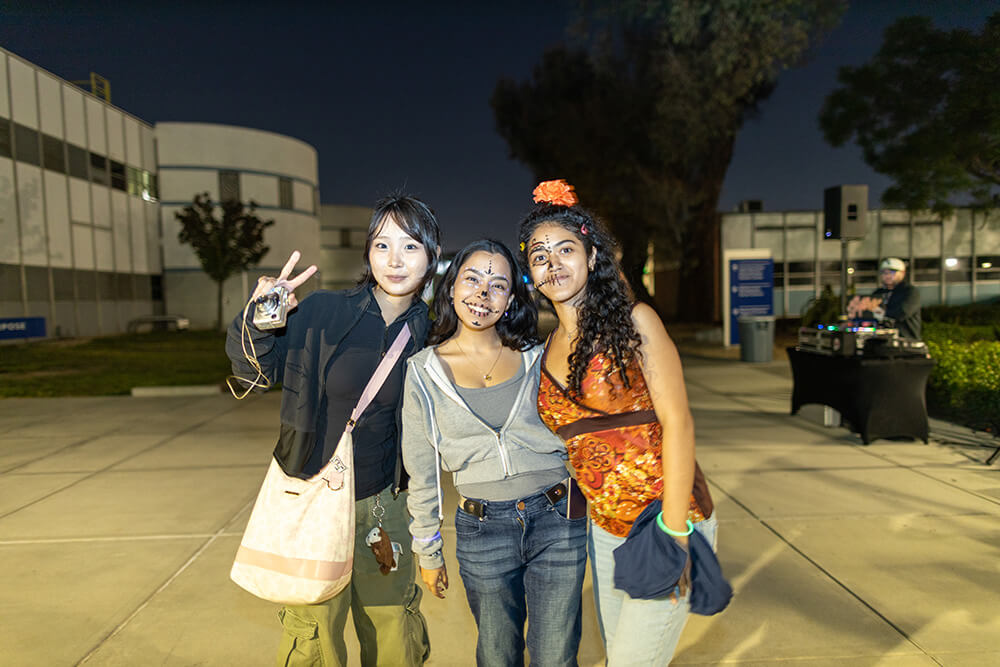 Three friends pose at night campus event, two with sugar skull face paint, one flashing a peace sign; DJ booth behind.