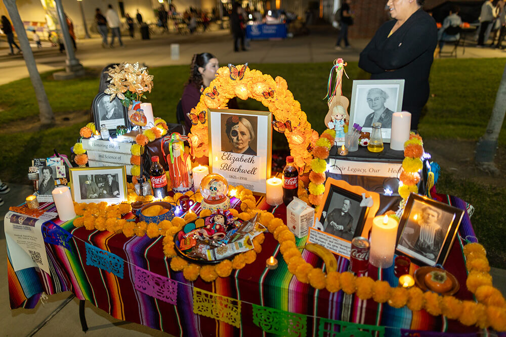 Altar honoring figures in science, centered on Elizabeth Blackwell portrait, with marigolds, candles, and sarape cloth.