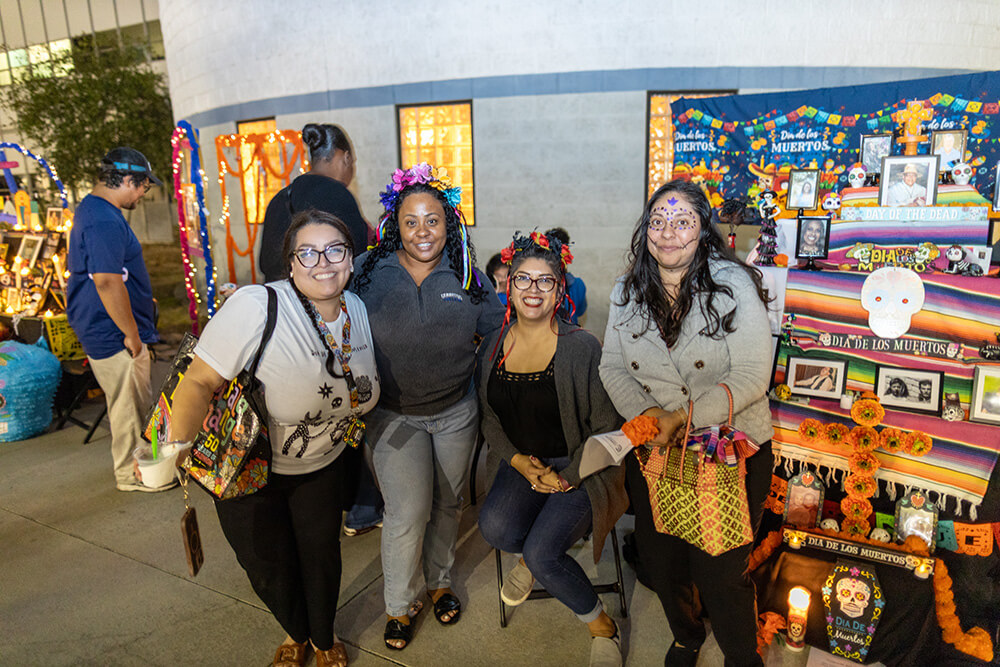 Four smiling women, some in flower crowns and face paint, pose beside a colorful Día de los Muertos altar with photos.