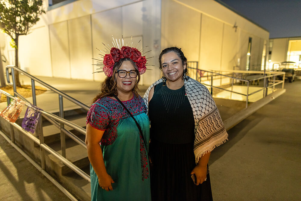 Two smiling women at night event, one in red rose crown and embroidered teal dress, the other in black dress and keffiyeh.