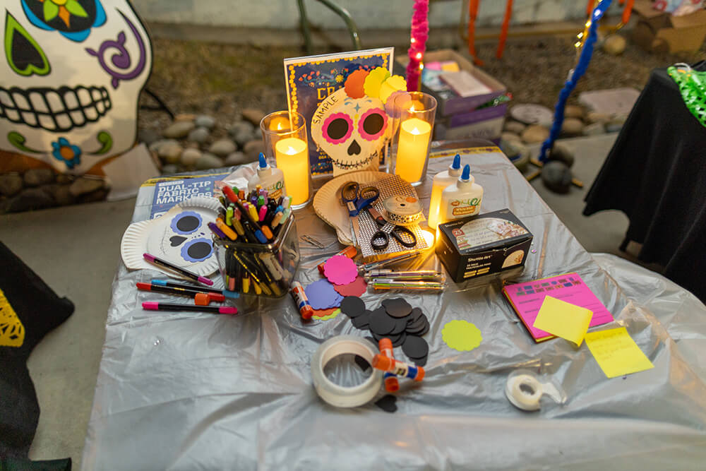 Craft table with paper plate sugar skull mask samples, markers, glue, scissors, and paper cutouts beside candles.