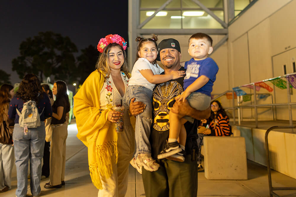 Family smiling at night festival: woman in flower crown and yellow shawl, man holding two kids, papel picado behind.