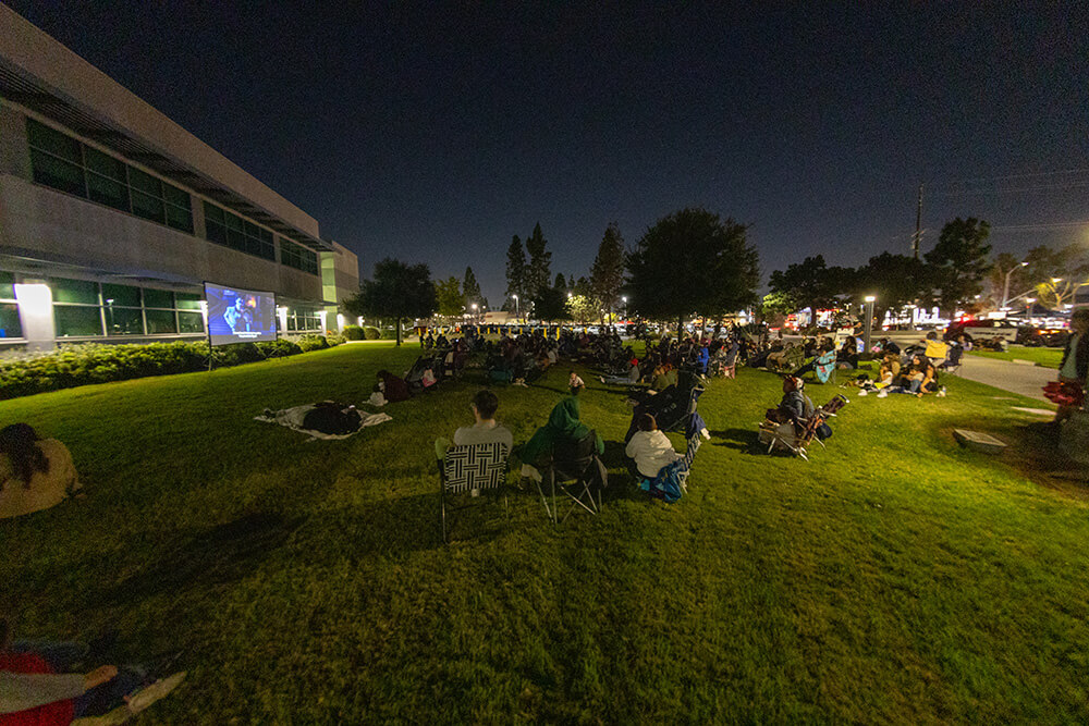 Crowd on lawn chairs and blankets watches an outdoor movie on a big screen beside a campus building at night.