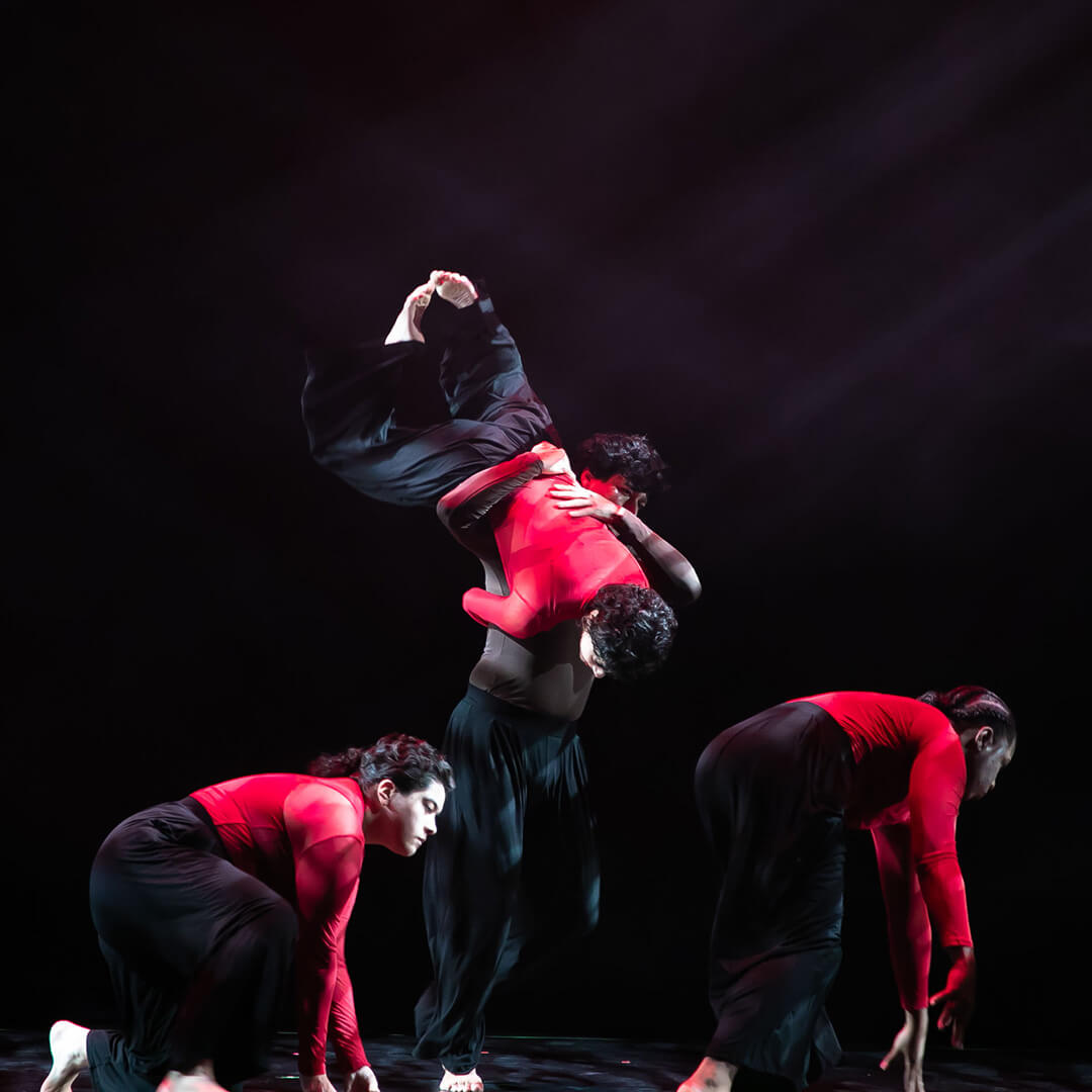 Four modern dancers in red and black perform a dynamic lift on a dark stage.