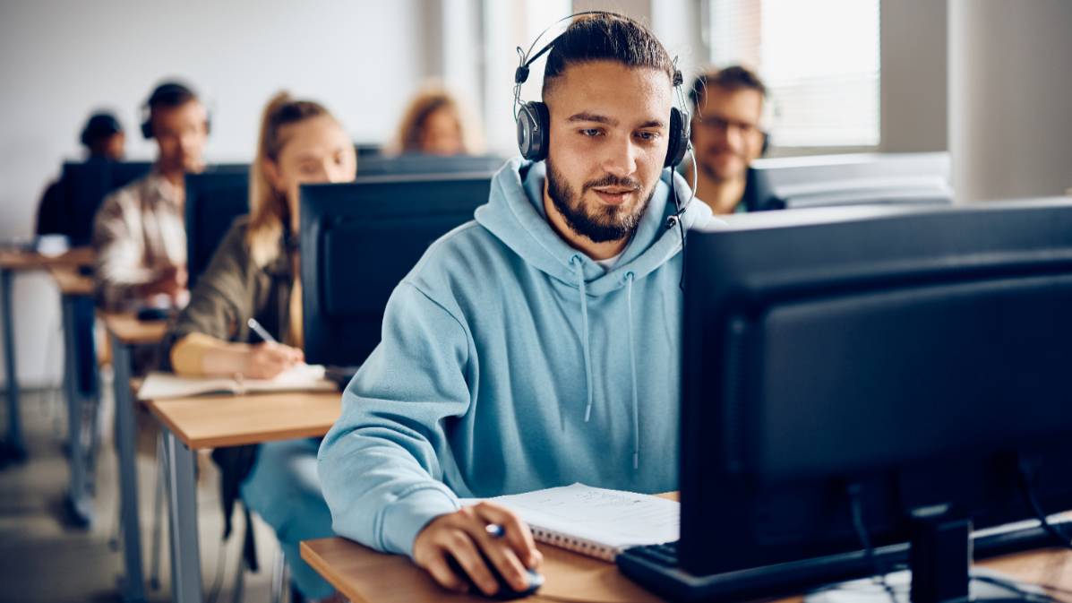 students sitting at desks working at computers while wearing headphones.