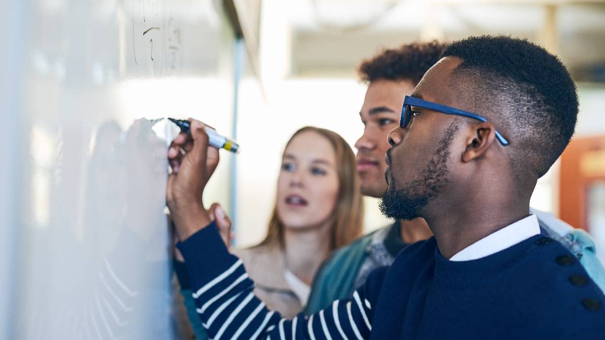 Three students looking at a whiteboard while one student writes on it.